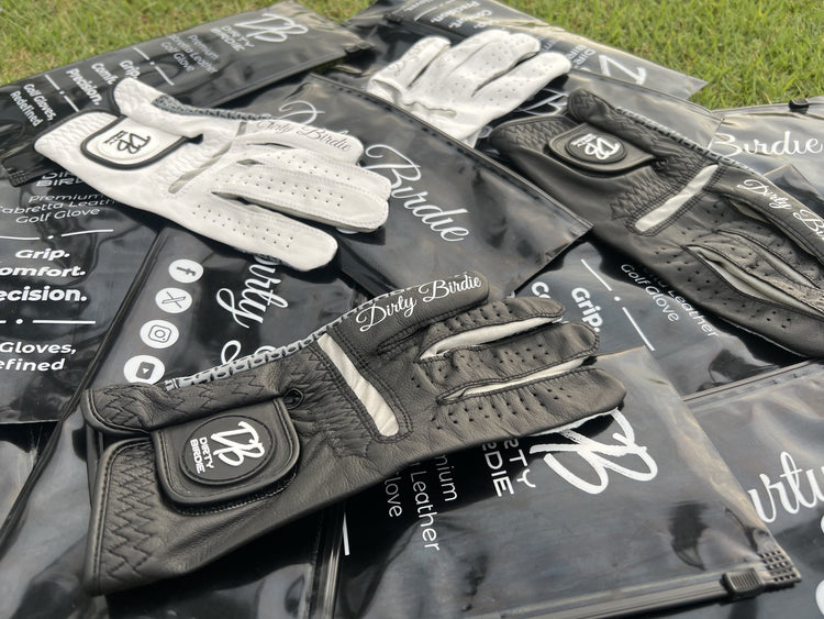 Black and white golf gloves laid on matching Dirty Birdie packaging, styled on fairway under the sun – premium cabretta leather with signature detailing