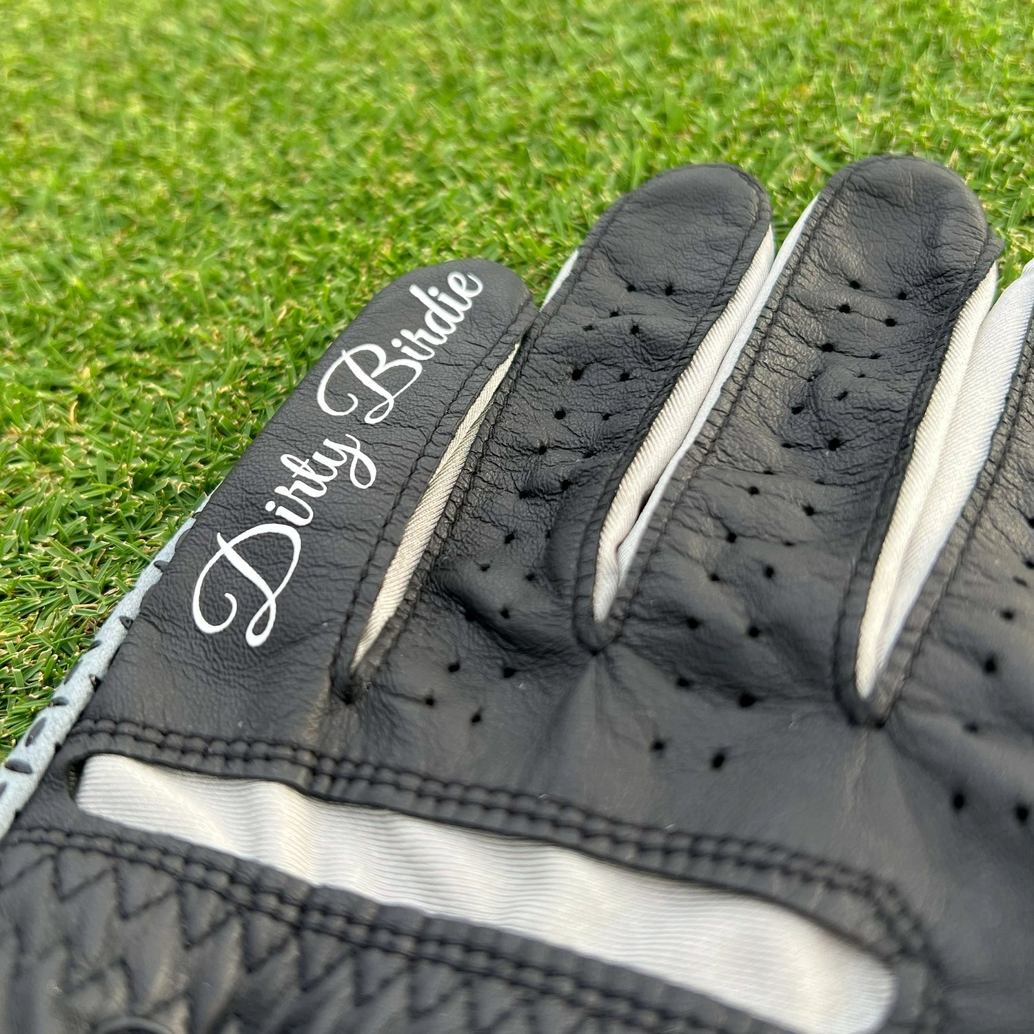 Black leather golf glove with Dirty Birdie branding on finger, close-up on grass background.