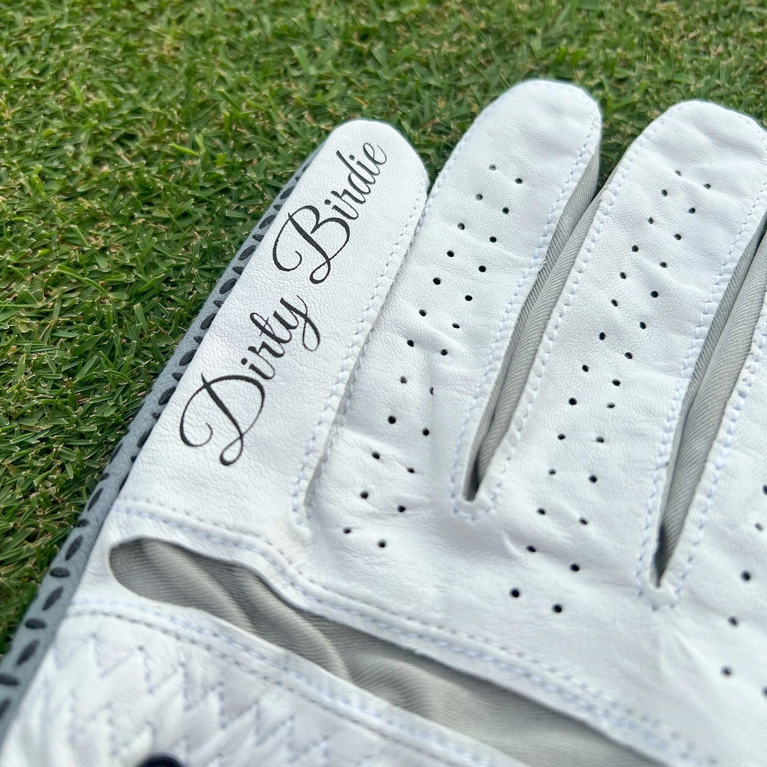 Detailed view of white golf glove showing Dirty Birdie branding on finger, laid on grass.